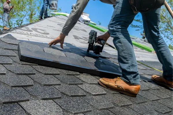 Roofer applying shingles with a nail gun and a few sheets of shingles beside him.